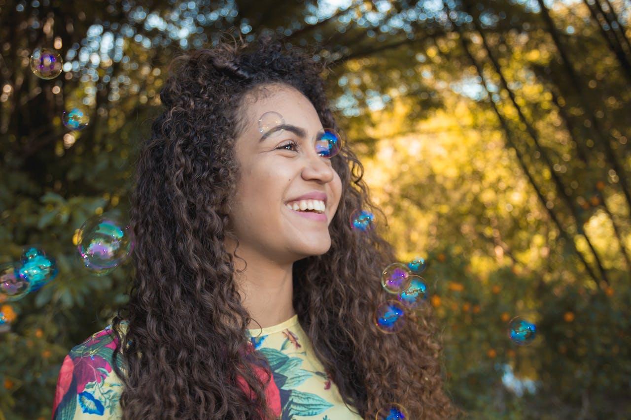 Joyful woman with curly hair enjoying colorful bubbles in a sunny outdoor setting.
