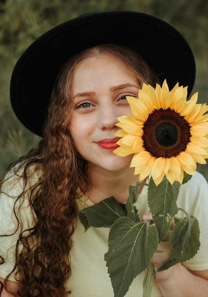 A smiling young woman holds a sunflower while wearing a hat, capturing a warm summer vibe.