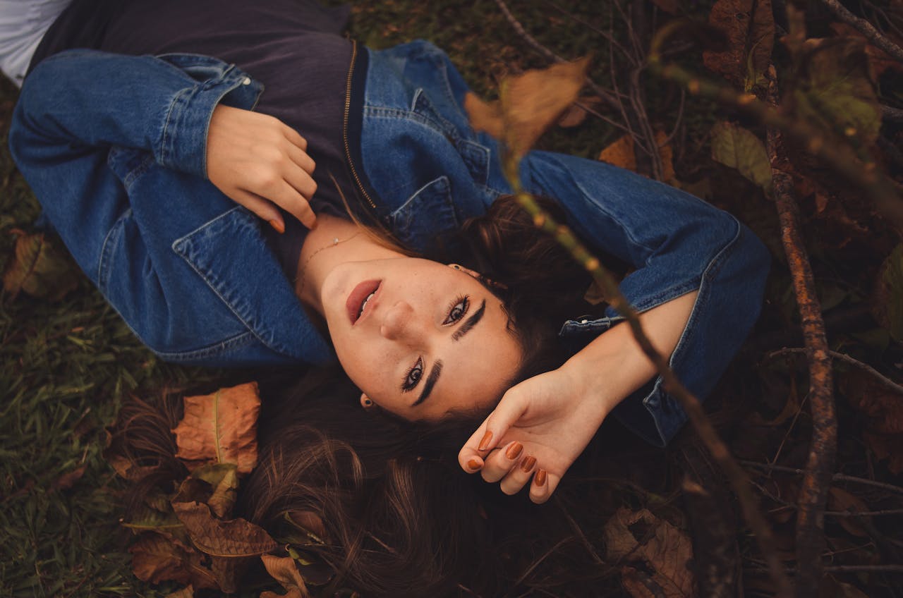 Young woman in a denim jacket posing on fallen autumn leaves, showcasing a serene and contemplative expression.